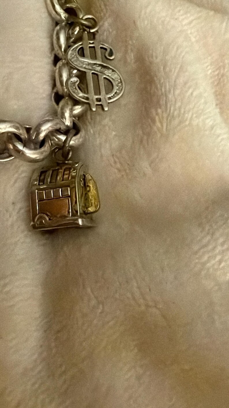 Puede incluir: Pulsera de plata con un charm de signo de d&oacute;lar y un peque&ntilde;o charm de tranv&iacute;a detallado. El charm del tranv&iacute;a tiene detalles dorados y cobrizos, a&ntilde;adiendo un toque de color a la cadena de plata. Fondo suave.