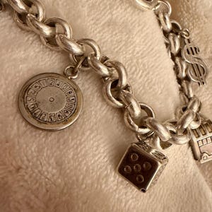 Puede incluir: Pulsera de plata con varios dijes, incluyendo un dado, una rueda estilo casino, un signo de d&oacute;lar y un autob&uacute;s de dos pisos. La pulsera tiene un dise&ntilde;o de cadena gruesa y se muestra sobre una superficie blanquecina.