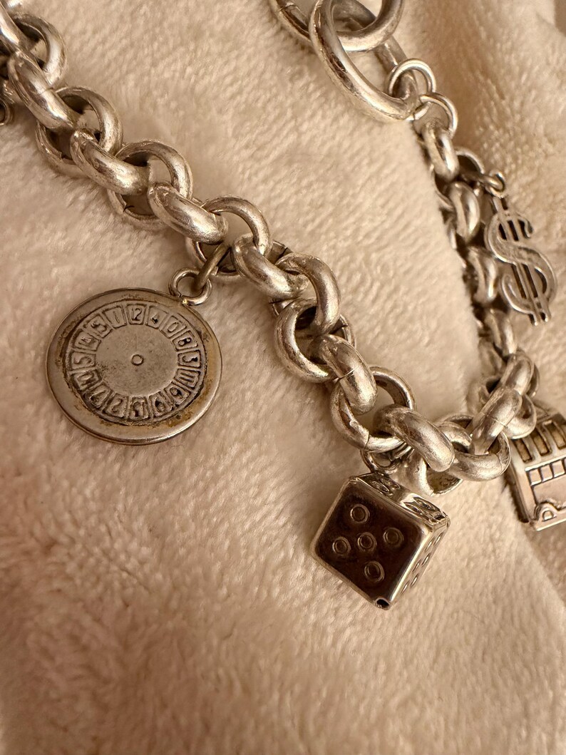 Puede incluir: Pulsera de plata con varios dijes, incluyendo un dado, una rueda estilo casino, un signo de d&oacute;lar y un autob&uacute;s de dos pisos. La pulsera tiene un dise&ntilde;o de cadena gruesa y se muestra sobre una superficie blanquecina.