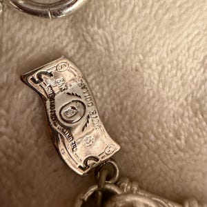 Puede incluir: Un dije plateado con forma de billete de cinco d&oacute;lares, con grabados detallados. El dije est&aacute; unido a una cadena, sobre un fondo suave y texturizado.