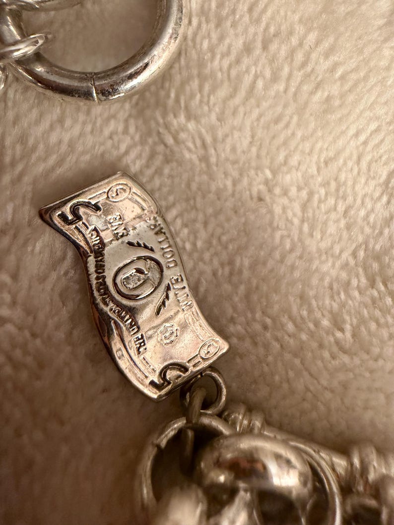 Puede incluir: Un dije plateado con forma de billete de cinco d&oacute;lares, con grabados detallados. El dije est&aacute; unido a una cadena, sobre un fondo suave y texturizado.