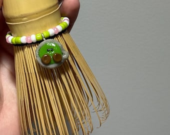 Könnte beinhalten: Ein Matcha-Besen aus Bambus mit einem dekorativen Perlenband in Rosa, Grün und Weiß. Eine kleine, grüne, cartoonartige Figur ist am Besen befestigt. Der Besen hat eine hellbraune Farbe.