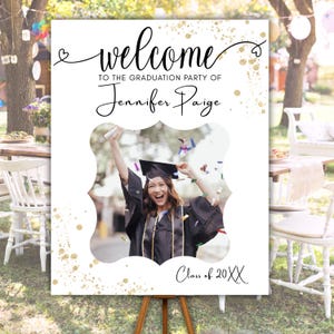 May include: A white graduation party welcome sign on a wooden easel. The sign has the words "welcome" and "To the graduation party of Jennifer Paige" in elegant script. A photo of a graduate is in the center, with the words "Class of 20XX" at the bottom.