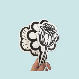 May include: A collection of die-cut stickers featuring black and white line art designs. The stickers include a detailed rose, a stylized flower with rounded petals, and other floral-inspired shapes with bold outlines and white borders.