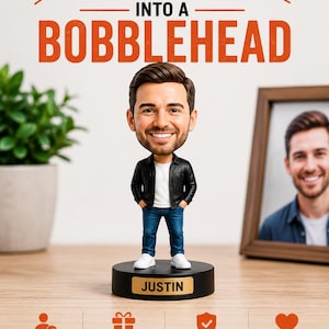May include: A custom bobblehead figure of a smiling person with brown hair, wearing a black jacket, white t-shirt, blue jeans, and white sneakers. The figure stands on a black circular base with the name JUSTIN printed in black on a gold plaque.