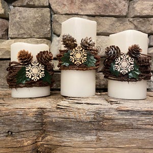 May include: Three white pillar candles decorated with pine cones, wooden snowflakes, and greenery. The candles are wrapped with a brown, twig-like material, creating a rustic, wintery aesthetic. The candles are arranged on a wooden surface.