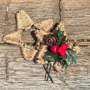 May include: A rustic Christmas hair clip featuring a star shape crafted from twine. It is adorned with a cluster of red berries, green holly leaves, and a small pine cone. The clip has black metal hair pins.