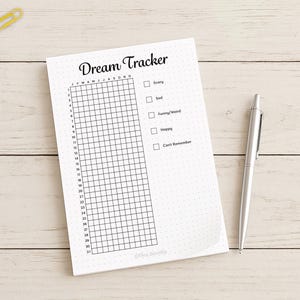 May include: A white notepad with the words "Dream Tracker" in black script. The notepad has a grid for tracking dreams, with checkboxes for emotions like "Scary" and "Happy." A silver pen rests beside the notepad. Colorful paperclips are scattered in the upper left corner.