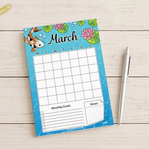 May include: A March calendar notepad with a koi fish and water lily design. The notepad has a blue background with a grid for daily entries, spaces for monthly goals and notes, and a silver pen.