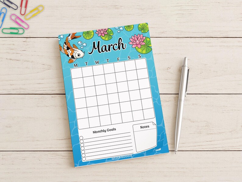 May include: A March calendar notepad with a koi fish and water lily design. The notepad has a blue background with a grid for daily entries, spaces for monthly goals and notes, and a silver pen.