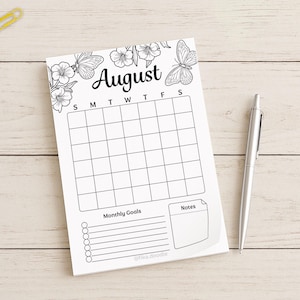 August monthly planner page with a blank calendar grid, monthly goals section, and notes area, photographed on a light wooden surface beside a pen and colorful paper clips.