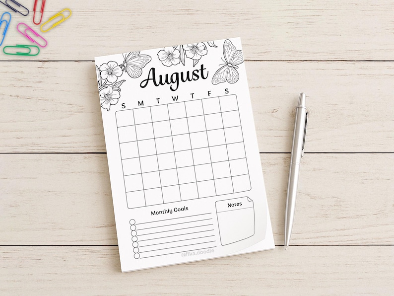 August monthly planner page with a blank calendar grid, monthly goals section, and notes area, photographed on a light wooden surface beside a pen and colorful paper clips.