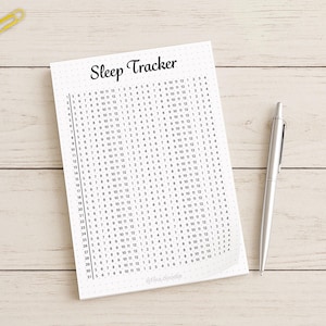 May include: A white sleep tracker journal with a grid of numbers, a silver pen, and colorful paper clips on a light wood surface. The journal's title, "Sleep Tracker," is written in black script.