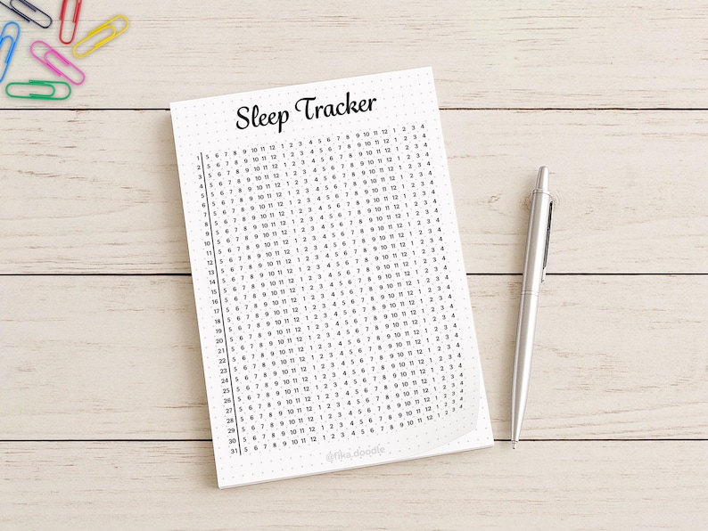May include: A white sleep tracker journal with a grid of numbers, a silver pen, and colorful paper clips on a light wood surface. The journal's title, "Sleep Tracker," is written in black script.