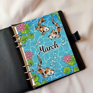 May include: A planner insert for March featuring three koi fish in a blue pond with lily pads and pink lotus flowers. The word "March" is written in a script font. The planner is black.