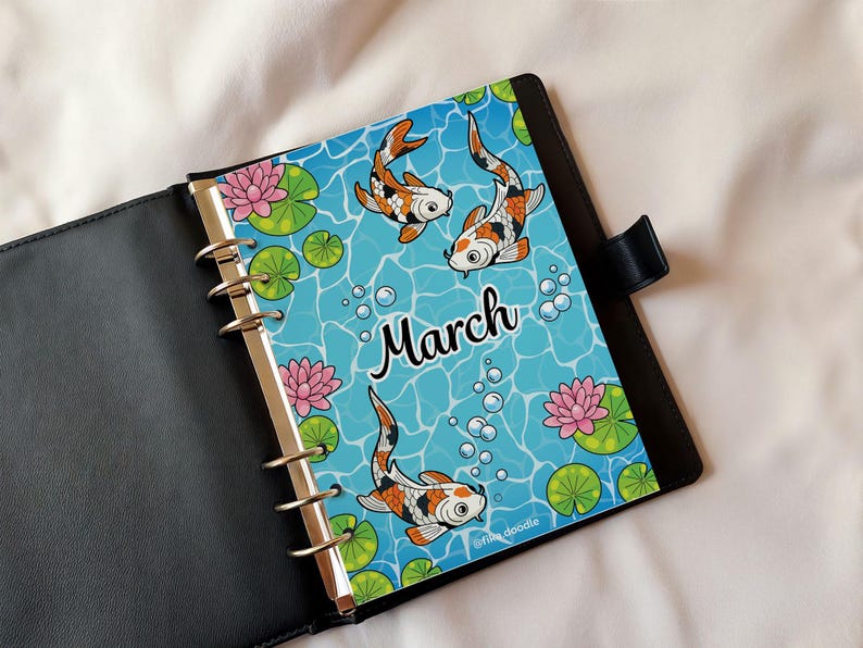 May include: A planner insert for March featuring three koi fish in a blue pond with lily pads and pink lotus flowers. The word "March" is written in a script font. The planner is black.
