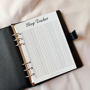 May include: Black leather binder with a sleep tracker insert. The insert is white with a dotted grid and a calendar-style layout for tracking sleep patterns. The words "Sleep Tracker" are at the top.