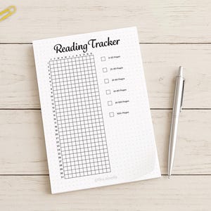 Op de afbeelding: Een witte "Reading Tracker" notitieblok met een raster om de leesvoortgang bij te houden. Het notitieblok heeft een titel en selectievakjes voor pagina-aantallen. Een zilveren pen ligt naast het notitieblok op een licht houten oppervlak.