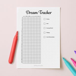 May include: A white Dream Tracker journal with a grid for daily entries and checkboxes for dream emotions. A red marker and three other colored markers are on a light pink surface.