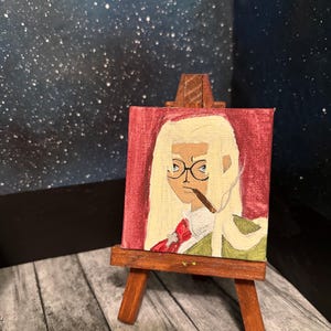 May include: A miniature painting on a small wooden easel. The artwork depicts a person with blonde hair, glasses, and a cigar, set against a red background. The person is wearing a green jacket and a red bow tie.