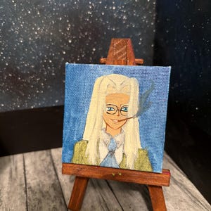 May include: Miniature painting on a wooden easel. The painting depicts a person with long blonde hair, glasses, and a cigarette. The background is blue, and the person is wearing a green and blue outfit. The easel is made of brown wood.