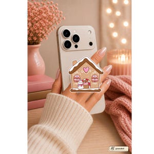 May include: A light-colored smartphone featuring a gingerbread house-shaped adhesive grip on the back. The grip displays a brown house with white icing details, a pink heart, two arched windows, and a small snowman figure standing by the door.