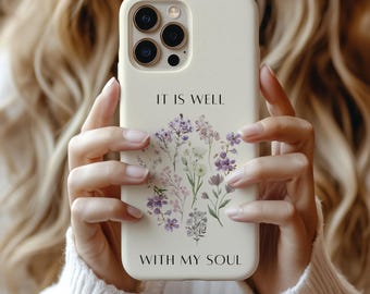 May include: A cream-colored phone case featuring the text "IT IS WELL WITH MY SOUL" above and below a floral design with purple and white flowers. The case has a camera cutout and is held by a person.