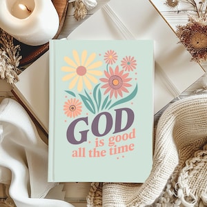 May include: A light green journal with a floral design featuring yellow, pink, and peach flowers. The words "GOD is good all the time" are printed in a mix of purple and pink fonts. The journal is open on a white surface.
