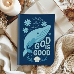 May include: A navy blue journal featuring a light blue whale illustration, flowers, and the text "GOD IS GOOD." The journal has a seashell and leaf design. The journal is open on a white surface.