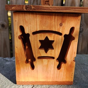 May include: Wooden box with a Star of David and scroll cutouts. The box is made of light brown wood and has a lid. A small wooden piece with Hebrew letters is on top of the box.