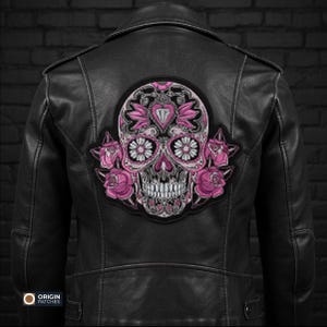Pode incluir: Jaqueta de couro preta com um grande patch bordado com um design de caveira de a&ccedil;&uacute;car rosa e branco. A caveira &eacute; decorada com motivos florais e de cora&ccedil;&atilde;o, e rodeada por rosas cor-de-rosa. A jaqueta tem um estilo motociclista cl&aacute;ssico.
