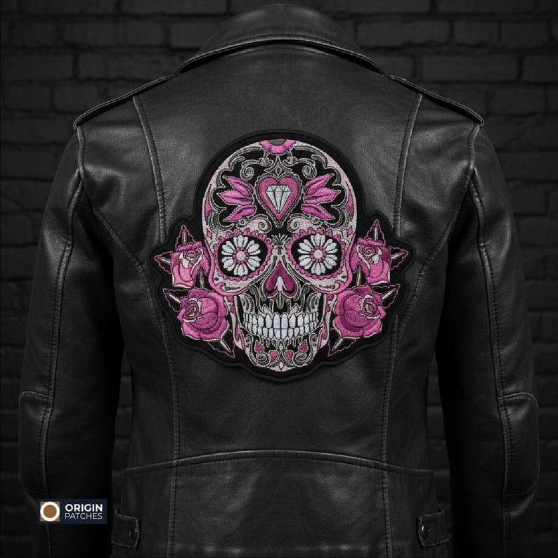Pode incluir: Jaqueta de couro preta com um grande patch bordado com um design de caveira de a&ccedil;&uacute;car rosa e branco. A caveira &eacute; decorada com motivos florais e de cora&ccedil;&atilde;o, e rodeada por rosas cor-de-rosa. A jaqueta tem um estilo motociclista cl&aacute;ssico.