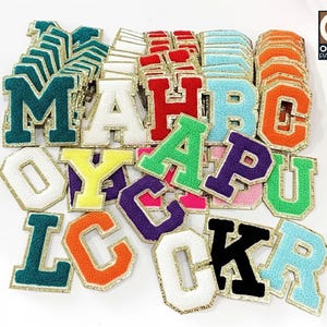 May include: A collection of varsity letter patches in various colors, including green, white, red, orange, purple, and blue. Each letter has a gold border. The letters are arranged on a white surface.