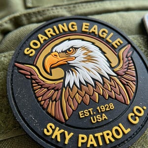 May include: A circular black patch featuring a raised graphic of a bald eagle head in white, brown, and orange. The text SOARING EAGLE is arched at the top, and SKY PATROL CO. is at the bottom. The text EST. 1928 USA is printed in the center.