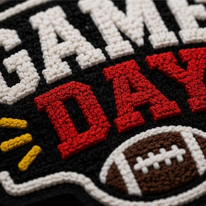 以下が含まれることがあります： 黒い背景に白と赤の立体的な刺繍でGAME DAYという文字が施されたテクスチャーのあるワッペン。デザインには3本の黄色いステッチラインと、白いレースが付いた茶色のフットボールが含まれており、全体が太い白い縁取りで囲まれています。