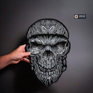 Pode incluir: Um grande patch de caveira preta com detalhes bordados brancos intrincados. O design apresenta uma caveira detalhada com uma express&atilde;o amea&ccedil;adora. O patch &eacute; exibido em um fundo cinza escuro, mostrando o artesanato.
