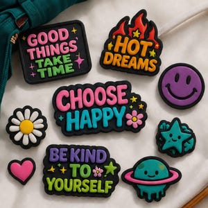 May include: A collection of colorful rubber shoe charms featuring various designs including a purple smiley face, a daisy, a pink heart, a teal star, a planet, and rectangular patches with the text Good Things Take Time, Hot Dreams, Choose Happy, and Be Kind To Yourself.