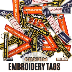 May include: A collection of custom embroidered tags in various colors, including orange, black, and navy blue. Many tags feature the text "REMOVE BEFORE FLIGHT" and "power stow". Some tags have an American flag design. The image also displays the text "CUSTOM EMBROIDERY TAGS".