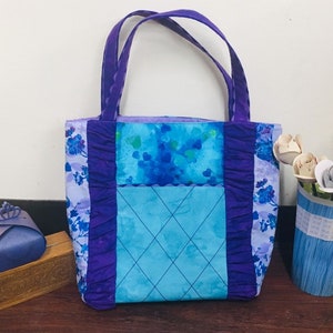 May include: A purple and blue tote bag with a floral print and a quilted pocket. The bag has a ruffled trim and purple handles.