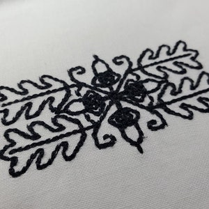 May include: Black embroidered design of stylized oak leaves and acorns on a white fabric background.