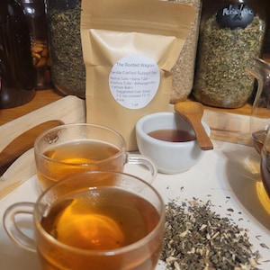 Puede incluir: Una presentaci&oacute;n de t&eacute; de hierbas. Dos tazas de vidrio transparente llenas de t&eacute; color &aacute;mbar est&aacute;n en primer plano. Un peque&ntilde;o cuenco blanco y una cuchara de madera est&aacute;n cerca. Tambi&eacute;n se ve una bolsa de papel marr&oacute;n y frascos de hierbas.