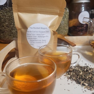 Puede incluir: Dos tazas de t&eacute; de vidrio transparente llenas de t&eacute; color &aacute;mbar se encuentran frente a una bolsa de papel marr&oacute;n. La bolsa est&aacute; etiquetada como "The Rooted Wagon" y enumera ingredientes como Rama Tulsi y Lemon Balm. Hierbas secas esparcidas cerca.