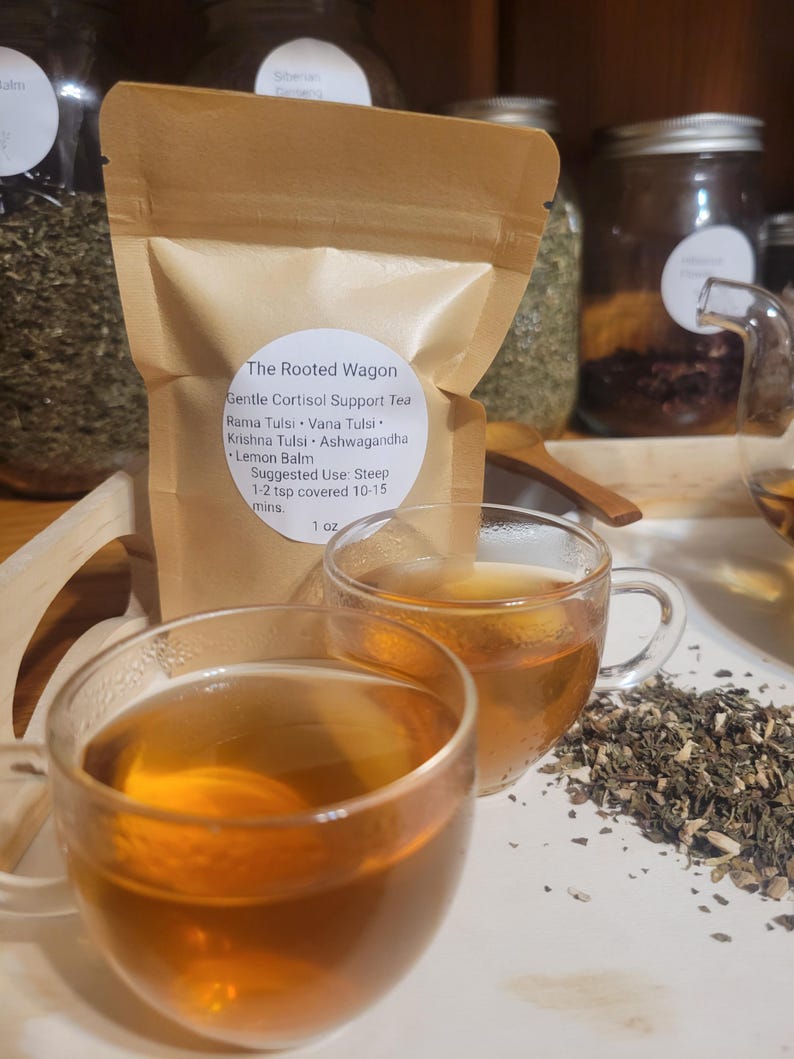 Puede incluir: Dos tazas de t&eacute; de vidrio transparente llenas de t&eacute; color &aacute;mbar se encuentran frente a una bolsa de papel marr&oacute;n. La bolsa est&aacute; etiquetada como "The Rooted Wagon" y enumera ingredientes como Rama Tulsi y Lemon Balm. Hierbas secas esparcidas cerca.