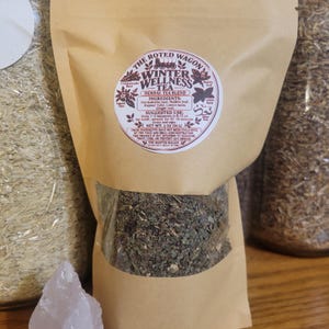 May include: A tan paper pouch containing a herbal tea blend labeled The Rooted Wagon Winter Wellness Tea. The label lists ingredients including Umckaloabo root, Mullein leaf, Kapoor Tulsi, Lemon balm, and Ginger root, with a net weight of 2 oz.