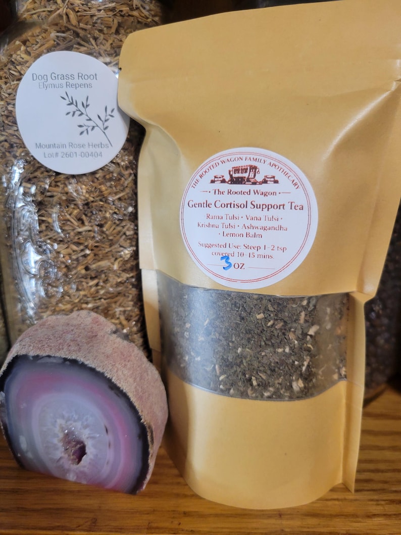 Puede incluir: Un paquete de "Gentle Cortisol Support Tea" de The Rooted Wagon Family Apothecary, con ingredientes como Rama Tulsi y Melisa. El paquete es de color beige con una ventana transparente, que muestra el t&eacute; en su interior. Una geoda cortada con tonos rosas y morados est&aacute; en primer plano.