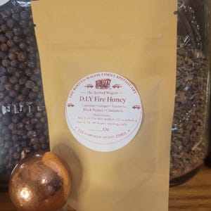 May include: A tan pouch labeled "D.I.Y Fire Honey" with a round sticker featuring text and illustrations. The ingredients listed are cayenne, ginger, turmeric, black pepper, and cinnamon. A copper-colored sphere sits in front of the pouch.