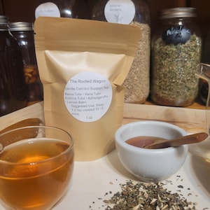 Puede incluir: Una exhibici&oacute;n de productos de t&eacute; de hierbas. Una bolsa de papel marr&oacute;n con la etiqueta "The Rooted Wagon" se encuentra junto a un vaso de t&eacute;, un cuenco peque&ntilde;o con una cuchara de madera y un mont&oacute;n de hierbas sueltas. Frascos de hierbas en el fondo.