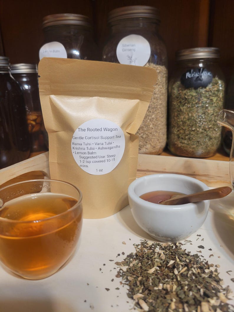 Puede incluir: Una exhibici&oacute;n de productos de t&eacute; de hierbas. Una bolsa de papel marr&oacute;n con la etiqueta "The Rooted Wagon" se encuentra junto a un vaso de t&eacute;, un cuenco peque&ntilde;o con una cuchara de madera y un mont&oacute;n de hierbas sueltas. Frascos de hierbas en el fondo.