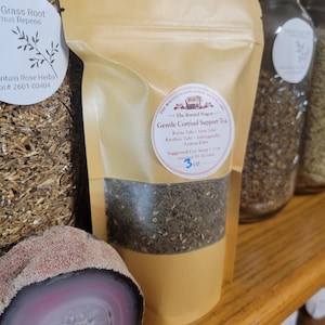 Puede incluir: Una bolsa de t&eacute; de color canela con la etiqueta "Gentle Cortisol Support Tea" con una ventana transparente que muestra las hojas de t&eacute;. La etiqueta incluye los ingredientes e instrucciones. Otros frascos de hierbas y una piedra decorativa est&aacute;n en el fondo.