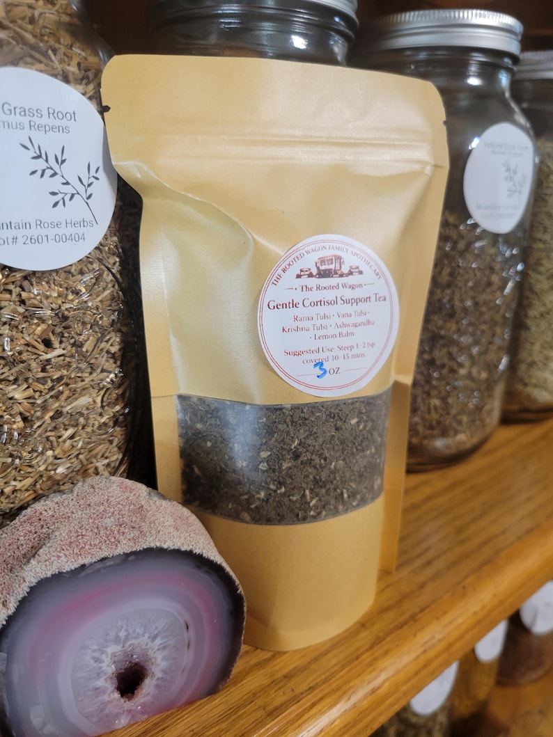 Puede incluir: Una bolsa de t&eacute; de color canela con la etiqueta "Gentle Cortisol Support Tea" con una ventana transparente que muestra las hojas de t&eacute;. La etiqueta incluye los ingredientes e instrucciones. Otros frascos de hierbas y una piedra decorativa est&aacute;n en el fondo.